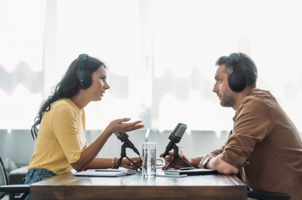 A man and a woman talking on a podcast episode. Looks like they're recording live and in a casual but engaging conversation that they're both enjoying. They're both wearing headphones and she's wearing a lovely yellow top while he's dressed in earth tones.