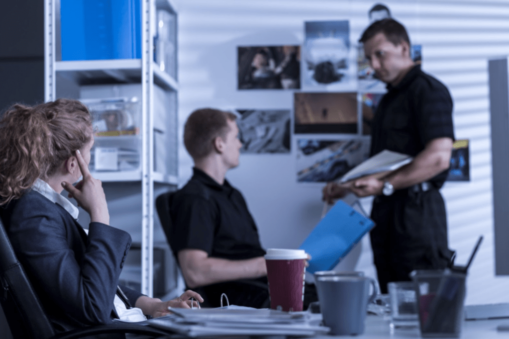 A team of opposition researchers meet in their shared office to discuss the specifics of their case. It's a male standing at the front of the room with two team members, a male and female, listening and contributing.