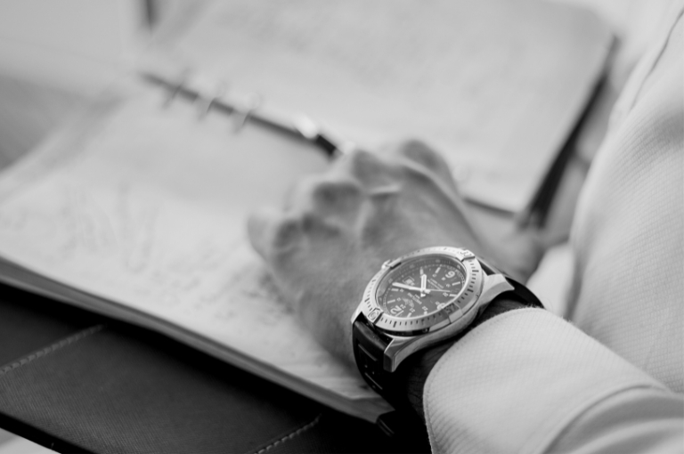A man is wearing a Breitling wrist watch while journaling in a leather bound notebook. Reveille Advisors works with high-net-worth individuals, family offices, and private equity with executive consulting.