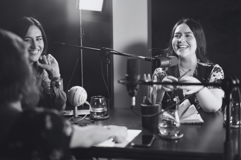 Three women sit around a studio table with microphones in front of them as they record a podcast episode. Reveille Advisors provides Strategic PR and Reputation Management services to clients in various roles.
